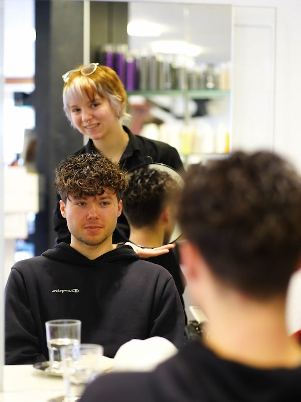Lächelnde Jungstylistin hält einem jungen Mann den Spiegel vor und zeigt den frisch geschnittenen Haarschnitt und die Dauerwelle von hinten