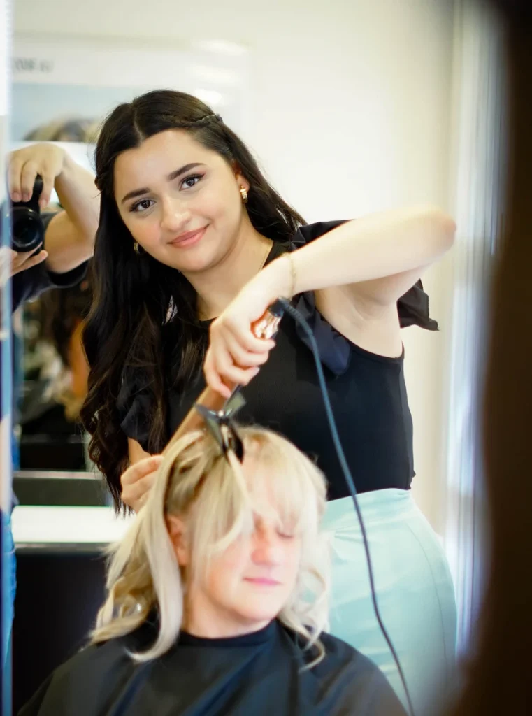 Junge Stylistin lächelt in die Kamera während sie einer Kundin mit dem Lockenstab Wellen in die Haare macht